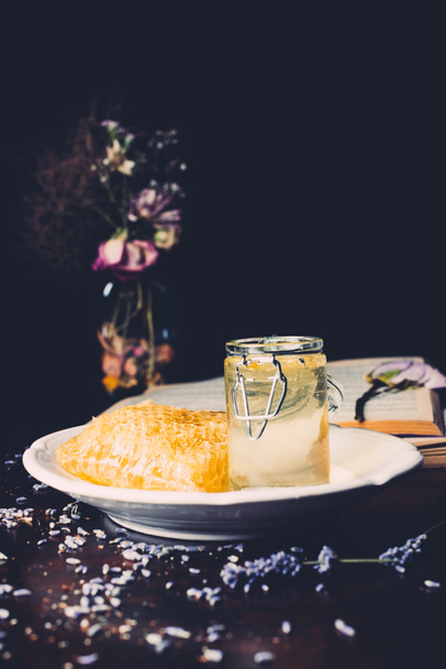 close up view of honeycomb and jar with honey surrounded by petals on black background  - Photo, Image
