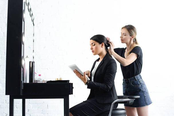 вид сбоку парикмахер делает прическу в то время как деловая женщина в костюме с помощью цифрового планшета
 - Фото, изображение