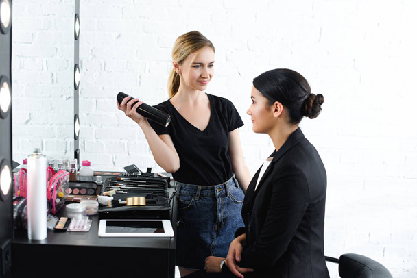 smiling hairstylist with hair spray and businesswoman in black suit - Photo, Image