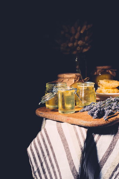 selective focus of cutting board with different jars of honey and lavender on black background - 写真・画像