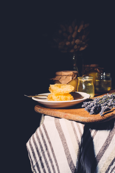 selektiver Fokus des Tellers mit Waben, Gläser mit Honig und Lavendel auf schwarzem Hintergrund - Foto, Bild