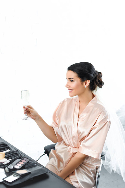 side view of smiling bride in silk bathrobe and veil with glass of champagne sitting at tabletop with cosmetics - Photo, Image