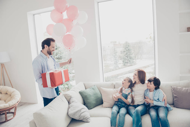 Portrait of husband prepare gift case in package for wife two kids holding air ballons, lovely mama sitting on couch hugging children meeting daddy with excited emotions clapping hands enjoying holiday - Foto, Imagem