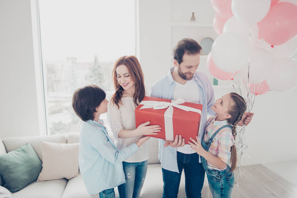 Portrait of attractive festive family having packed box in red package with bow air ballons looking at each other mum daddy two kids enjoying event indoor. Good mood atmosphere idyllic concept - Photo, Image