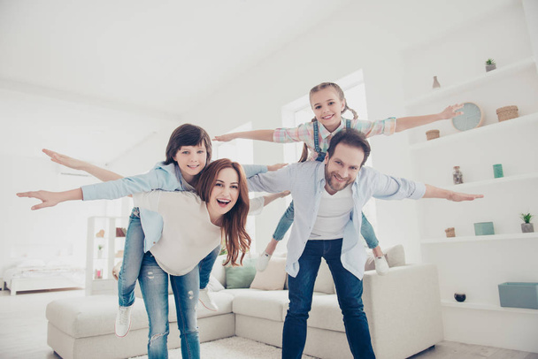 Low angle bottom view of creative joyful family, parents holding on back in piggyback style two kids open hands to the side making airplane in modern white room. Aspiration imagination concept - Fotó, kép