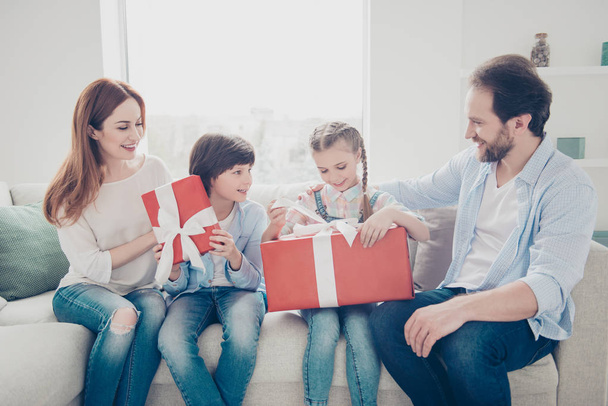 Portrait of careful family unpacking gift cases in red package with bow together sitting on sofa indoor enjoying surprise holding presents in hands speaking talking having fun - Foto, Bild