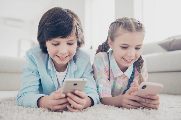 Portrait of two kids lying on stomach on floor carpet holding smart phones in hands using wifi 3G internet enjoying weekend resting indoor chatting with parents texting sms - Foto, immagini