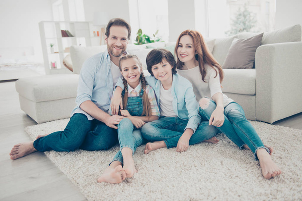 Portrait of lovely cheerful family with two kids in casual outfits sitting on carpet near sofa in modern white livingroom looking at camera. Domicile domestic lifestyle rest relax leisure concept - Photo, Image