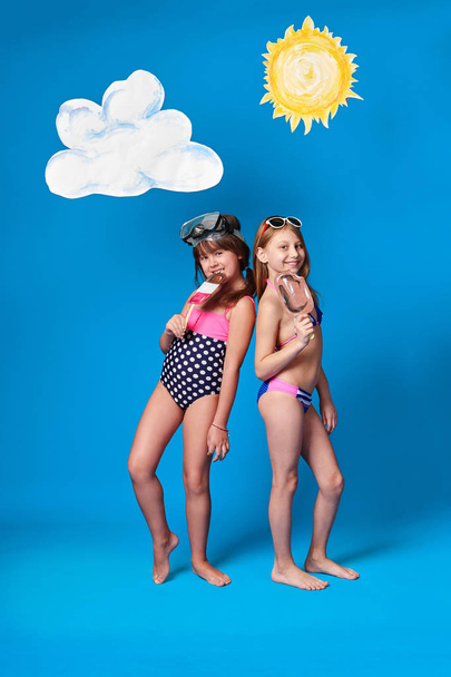 Concept summer,beach.Children girls model colorful swimsuit posing,holding hand ice cream,on head mask diving. Caucasian kids,blue background with sun,cloud in studio. Advertising,sea recreation. - Фото, изображение