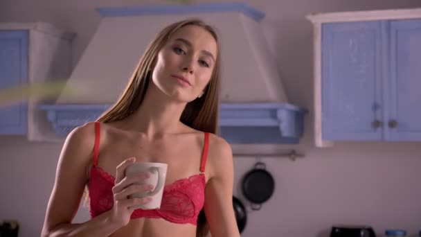 Young sexy woman standing in kitchen and holding cup of coffee, smiling at camera, cute and attractive - Video, Çekim