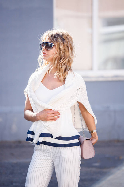 Outdoor portrait of beauty blond model wearing stylish white clothes and sunglasses. Young woman standing on street at daytime. - Φωτογραφία, εικόνα