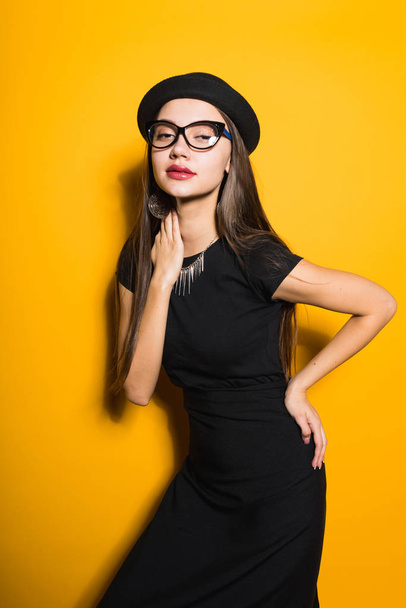 luxurious stylish girl model in a trendy black hat, dress and glasses posing on a yellow background - Foto, Imagen