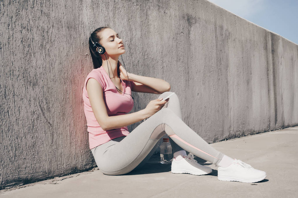 mujer joven y tranquila en ropa deportiva sentada en el suelo, descansando después del entrenamiento y escuchando música en los auriculares
 - Foto, imagen