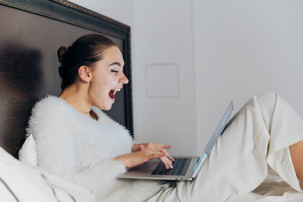 joyful laughing girl in white pajamas sitting in bed late at night, watching funny video on her laptop - Foto, imagen