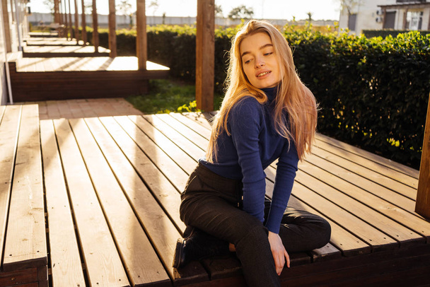 attractive young blond model girl in fashionable blue sweater sits on a wooden bench in the park, smiling - Foto, Bild
