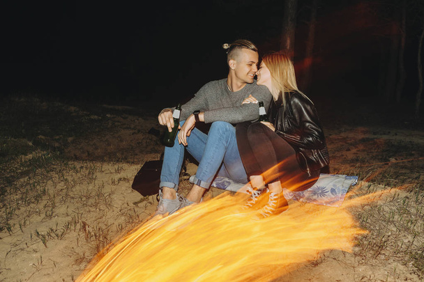 Handsome man and beautiful woman with bottles of beer sitting near burning fire and touching noses in evening - Fotoğraf, Görsel