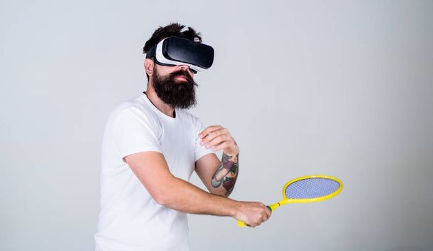 Man with beard in VR glasses beating pitch, grey background. Guy with VR glasses play tennis with racket. Virtual sport concept. Hipster on busy face use modern technology for sport games - Foto, afbeelding