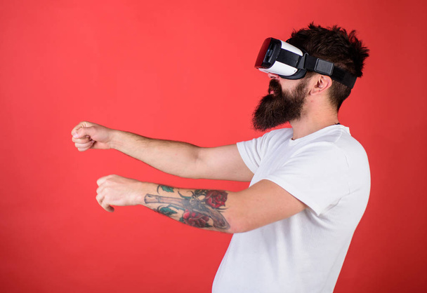 Hipster on confident face driving bike in virtual reality with modern digital gadget. Man with beard in VR glasses driving motorbike, red background. Virtual driving lessons concept - Photo, image