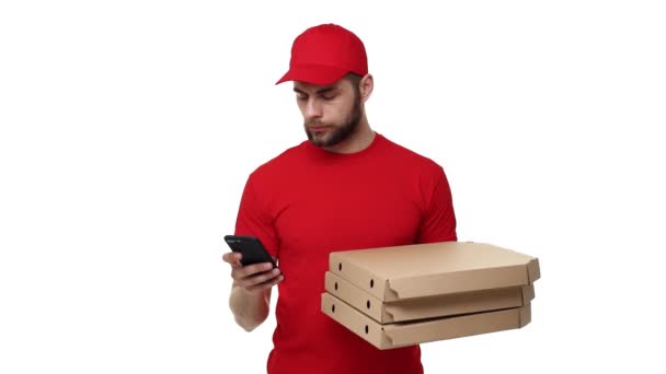 Delivery boy in a red uniform holding a stack of pizza boxes and checking order on mobile phone. Isolated over white background. - Footage, Video