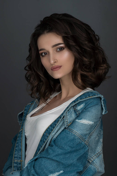 beautiful girl fashion model with hairstyle and makeup poses positively at the camera in the studio on a gray background in a white mike and jeans. - 写真・画像