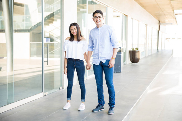 Longitud completa de hombre y mujer adolescentes sonrientes tomados de la mano mientras están de pie juntos en el pasillo del centro comercial
  - Foto, Imagen