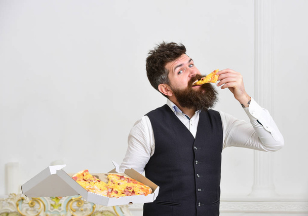 Pizza delivery concept. Macho in classic clothes hungry, holds slice of cheese pizza, eats, white background. Man with beard and mustache holds delivered box with tasty fresh hot pizza - Valokuva, kuva
