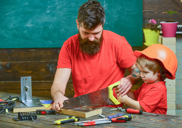 Masculine duties concept. Father, parent with beard teaching little son to sawing with sharp handsaw, carpenter crafts. Boy, child busy in protective helmet learning to use handsaw with dad - Foto, Imagen