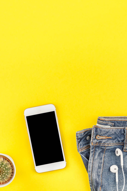 Flat lay of blue jeans and smartphone with headphones on bold yellow paper background with copy space. Overhead view of woman casual outfit. Trendy hipster look top view - Fotó, kép