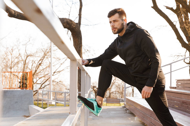 Portrait of confident young sportsman in earphones doing stretching exercises on steps outdoors - Фото, зображення