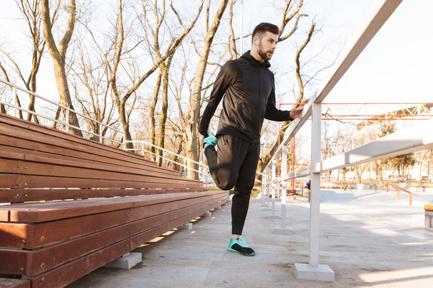 Portrait of young sportsman in earphones doing exercises outdoors - Zdjęcie, obraz
