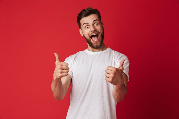Picture of happy young handsome man standing isolated over red wall background. Looking camera showing thumbs up. - Valokuva, kuva