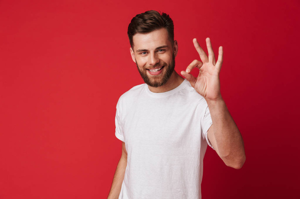 Picture of happy young handsome man standing isolated over red wall background. Looking camera showing okay gesture. - Fotografie, Obrázek