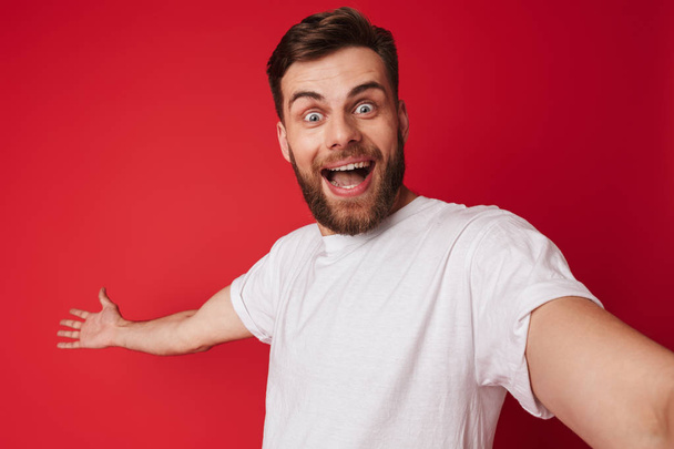 Picture of emotional excited young handsome man standing isolated over red wall background make a selfie looking camera. - Fotografie, Obrázek