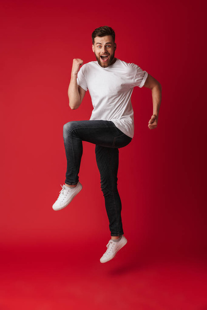 Photo of handsome young man jumping isolated over red wall background showing winner gesture. - Foto, Imagem