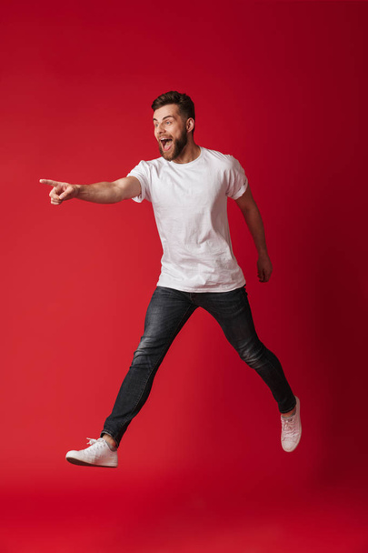 Photo of handsome young man jumping isolated over red wall background pointing looking aside. - Фото, зображення