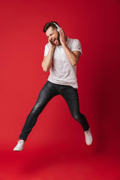 Image of emotional screaming young man isolated over red wall background listening music with headphones. - Foto, imagen