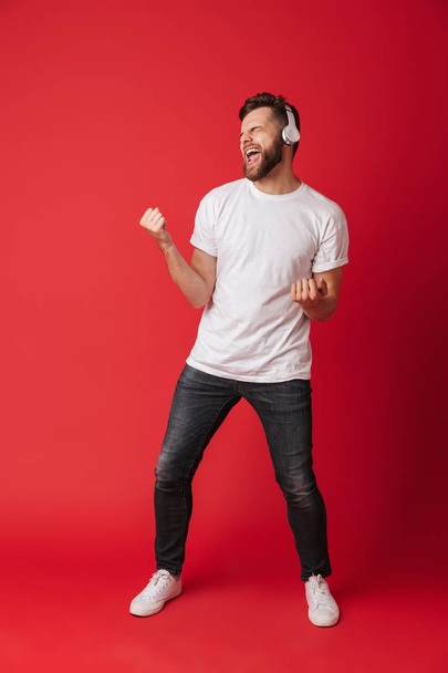 Image of emotional screaming young man isolated over red wall background listening music with headphones. - Fotoğraf, Görsel