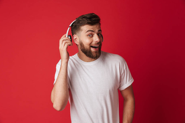 Foto van de knappe geschokt jongeman geïsoleerd over rode muur achtergrond luisteren muziek met koptelefoon. - Foto, afbeelding