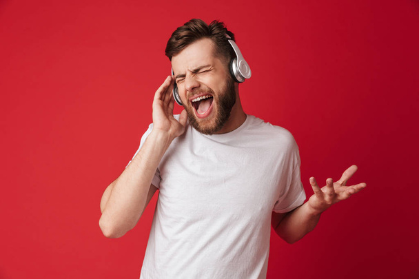Image of emotional screaming young man isolated over red wall background listening music with headphones. - Foto, immagini