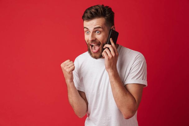 Photo of handsome young surprised man standing isolated over red wall background talking by mobile phone make winner gesture. - Foto, afbeelding