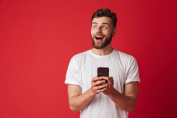 Photo of shocked young man standing isolated over red wall background using mobile phone. - Zdjęcie, obraz