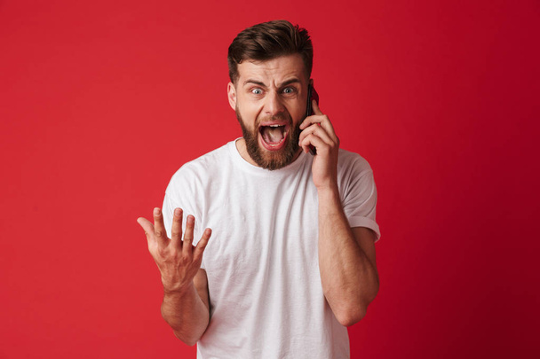 Image of screaming young man standing isolated over red wall background talking by mobile phone. - Photo, image