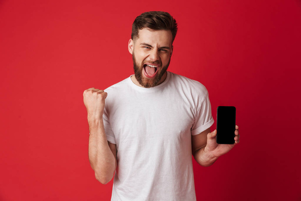 Image of excited young man standing isolated over red wall background showing display of mobile phone make winner gesture. - Fotoğraf, Görsel