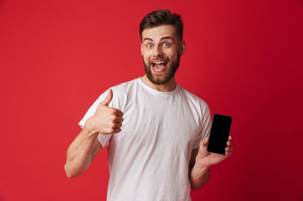 Image of happy young man standing isolated over red wall background showing display of mobile phone make thumbs up gesture. - 写真・画像