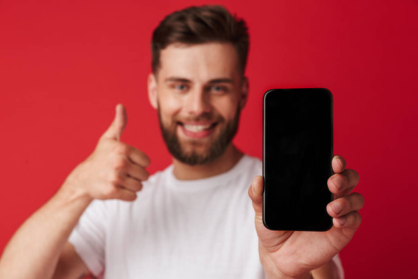 Image of happy young man standing isolated over red wall background showing display of mobile phone make thumbs up gesture. - Фото, зображення