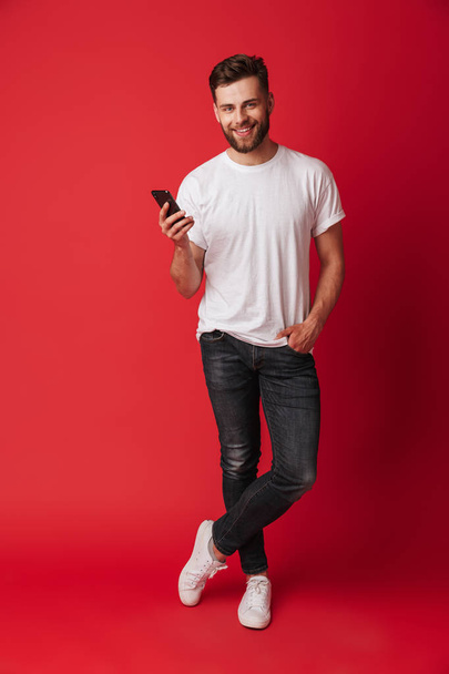 Image of happy young man standing isolated over red wall background using mobile phone. - Zdjęcie, obraz