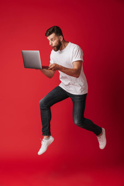 Εικόνα έκπληκτος νεαρός άνδρας άλματα απομονώνονται σε φόντο κόκκινο τοίχο χρησιμοποιώντας φορητό υπολογιστή. Κάμερα που αναζητούν. - Φωτογραφία, εικόνα
