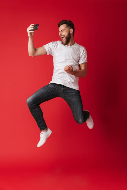 Image of excited young man isolated over red wall background make selfie by mobile phone jumping. - Foto, immagini