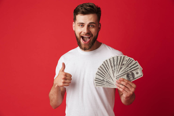 Image of happy young man isolated over red wall background holding money make thumbs up. - Foto, Bild