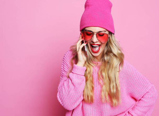 chica sonriente cool feliz hablando en el teléfono inteligente en ropa rosa sobre fondo rosa con gafas de sol rosa sombrero rosa y jersey rosa. Concepto de estilo de vida
. - Foto, Imagen